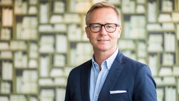 En man i kostym och glasögon står framför en vägg.