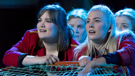 Två kvinnliga elever är med i en pjäs, har röda jackor på sig, tittar uppät. 