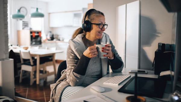 En leende kvinna sitter vid sin dator med en kopp kaffe i handen och samtalar med sin lärare på en distanskurs med Folkuniversitetet i Södertälje.