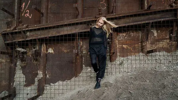 Kvinna i svarta kl&auml;der lutar sig mot en rostig v&auml;gg.