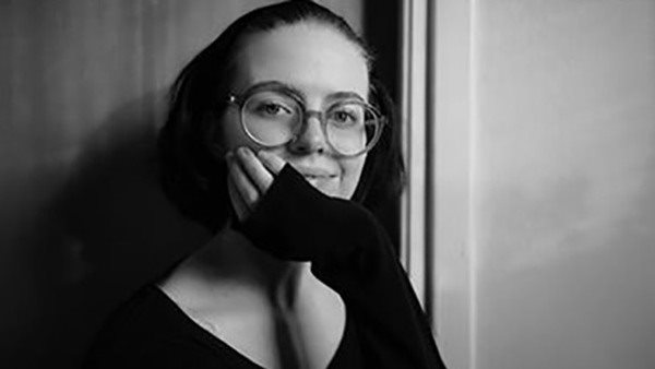 Svartvit porträttbild av Ellen Skarelius, elev i SA18 på Kungstensgymnasiet. Ellen har glasögon och ler, med hakan vilande i ena handen.