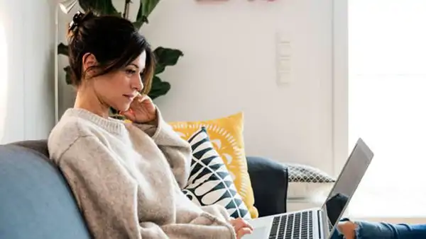 Kvinna sitter i soffa med laptop i knäet och deltar fokuserat i en distanskurs