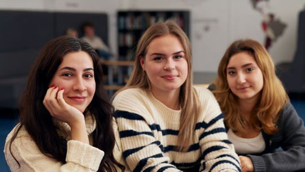 Tre elever på Internationella gymnasiets samlingsplats tittar in i kameran.