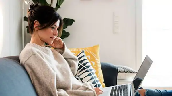 Kvinna sitter i soffa med laptop i knäet och deltar fokuserat i en distanskurs