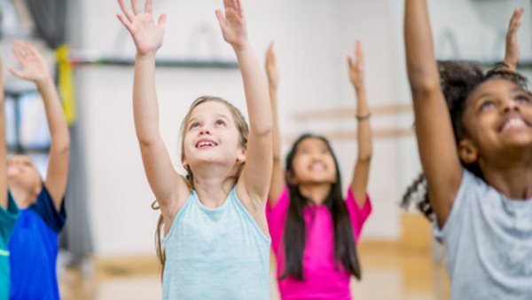 A group of elementary school children are indoors in a gym. They are wearing casual athletic clothing. They are sitting on yoga mats and raising their arms into the air.