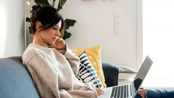 Kvinna sitter i soffan hemma med laptop i knäet och tittar på skärmen under en distanskurs.