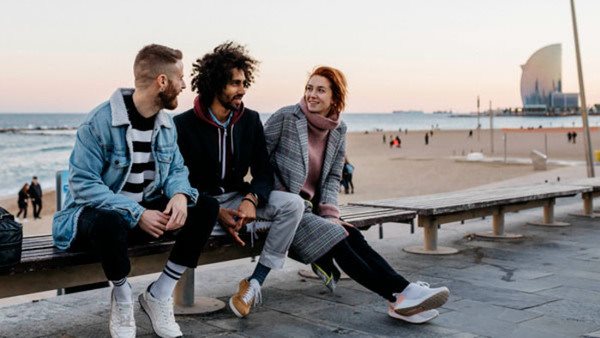 Tre unga vuxna skrattar på en bänk vid stranden i Barcelona - efter kurs i katalanska på Folkuniversitetet