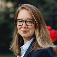 Cathrine Simons har lånt mörkblondt hår och glasögon och tittar sig lätt över axeln mot kameran.