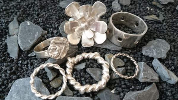 Handgjorda smycken i silver, som ringar och blomformad brosch, visas upp på mörk sten - kurs i silversmide