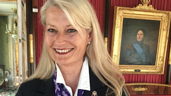 Porträttbild på Ulrica Häller i långt ljust hår och stort leende i slottsmiljö. På väggen bakom henne hänger ett kungaporträtt i guldram.