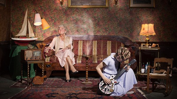 Äldre kvinna sitter i soffa och en flicka spelar elgitarr sittande på golvetmöblerat rum