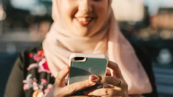 Leende person med ljusrosa hijab tittar på sin mobil utomhus