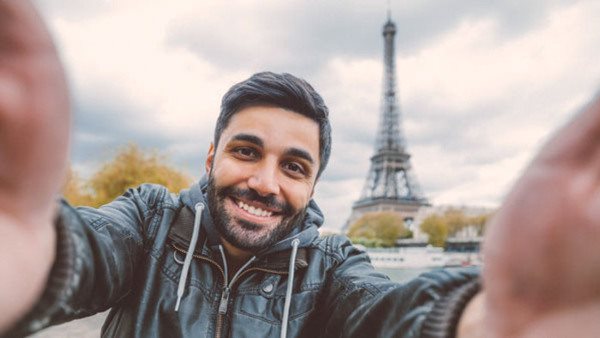 Young middle eastern man holding smart phone and taking selfie pictures. He is happy, smiling enjoying and looking at her camera at coastline of Seine river against Tour Eiffel in Paris, France.