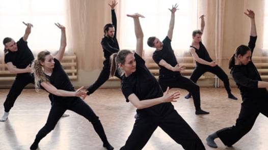 Three fit girls and four guys in black activewear standing on the floor with outstretched legs and bent knees while exercising in dance studio