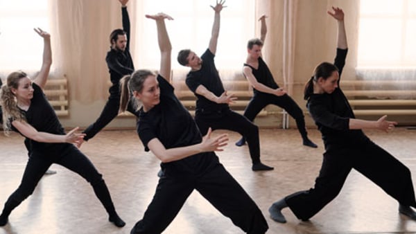 Three fit girls and four guys in black activewear standing on the floor with outstretched legs and bent knees while exercising in dance studio