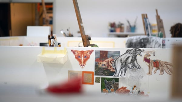 Ljus ateljé med flera studenter som arbetar vid skärmar och stafflier. Kreativ miljö med målningar och arbetsmaterial.