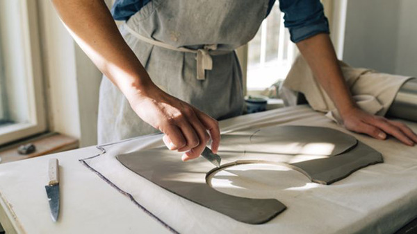 Närbild på händer som skär i lera vid ett bord där solens strålar lyser in.