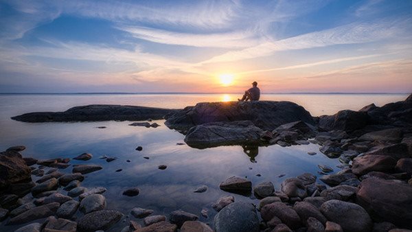 En man sitter på en klippa vid havet i solnedgågn.