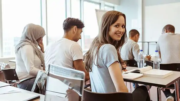 Fem personer i ett klassrum, där en av personerna en ung tjej med brunt långt hår vänder sig till kameran och ler.