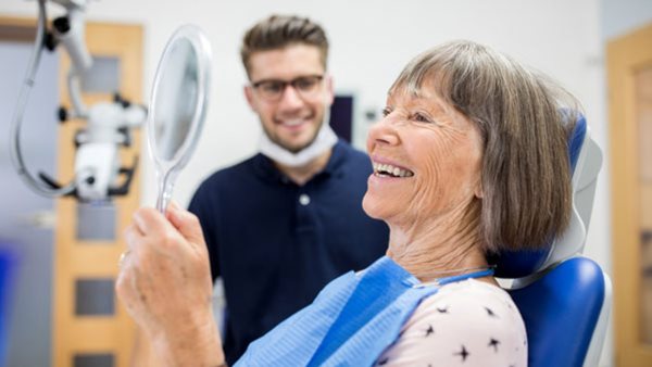 Smiling elderly patient checking her teeth into a hand mirror after her dental treatment. Senior patient checking her teeth after treatment at dental clinic.