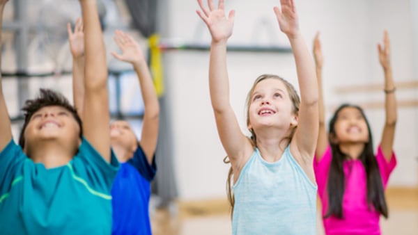 A group of elementary school children are indoors in a gym. They are wearing casual athletic clothing. They are sitting on yoga mats and raising their arms into the air.