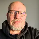 En man med skägg och glasögon, klädd i en svart huvtröja.