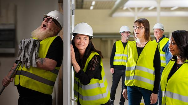 personer i hjälmar och varselväst lyssnar på person som sjunger bakom en dörr