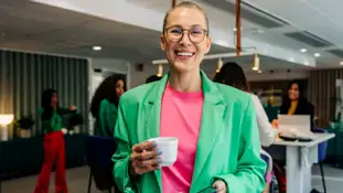 En kvinna i grön jacka med en kopp kaffe i handen under en kafferast på Folkuniversitetet