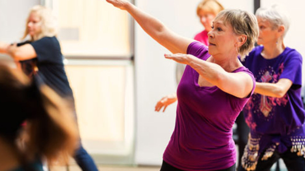 Diverse group of senior women take dance lessons in exercise studio at senior center. The women have their arms up as they learn the dance. Large bright windows are in the background.
