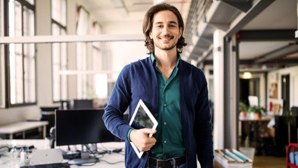 Portrait of smiling young handsome businessman standing with digital tablet at creative office