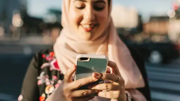 Leende person med ljusrosa hijab tittar på sin mobil utomhus