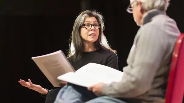En äldre kvinna och man spelar teater med manus i händerna. 
