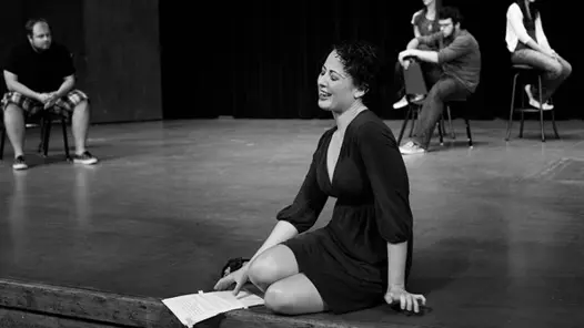 Kvinna i svart klänning sitter på en scen med manus i handen