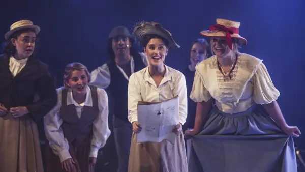 Tre elever på scen under en föreställning, iklädda i kläder från sen 1800-talet, med hattar och långa kjolar. 