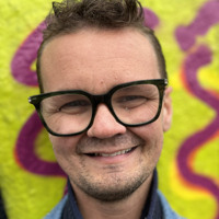 En man med glasögon står framför en graffitimålad vägg.