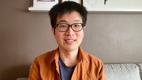 Porträtt av Tianle Yu, en asiatisk man med mörkt hår och glasögon. Tianle sitter i ett vardagsrum och har en orange skjorta på sig.