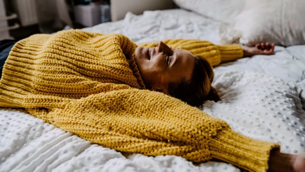 En kvinna i gul stickad tröja ligger i en säng med armarna utsträckta ovanför huvudet.