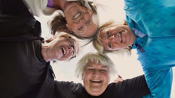 Fyra äldre kvinnor står i en cirkel och håller om varandra, medan de tittar ner i kameran och skrattar.