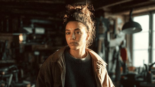 En kvinnlig student som står i en verkstad med brun jacka