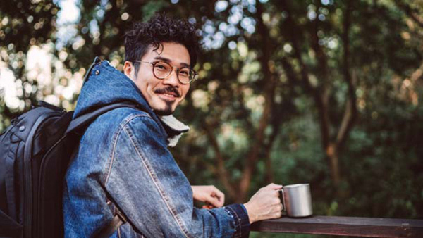 Young handsome asian man smiling joyfully at camera while drinking with his reusable coffee cup in country park