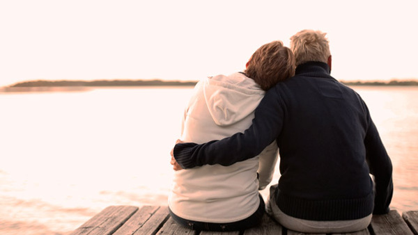 Två personer sitter på en brygga med ryggarna mot kameran. En håller om den andre som lutar sitt huvud mot dennes axel.