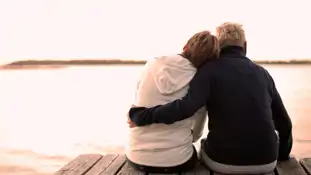 Två personer sitter på en brygga med ryggarna mot kameran. En håller om den andre som lutar sitt huvud mot dennes axel.
