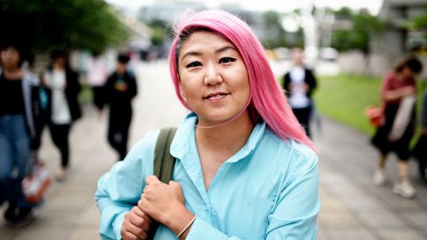 Ung person med rosa hår och blå skjorta ler mot kameran på en stadsgata - deltagare på en kurs i färöiska