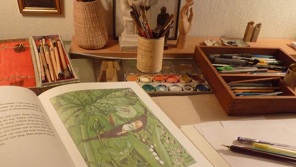 Arbetsbord med akvarellfärger, pennor och illustrerat bokuppslag under kurs i illustration