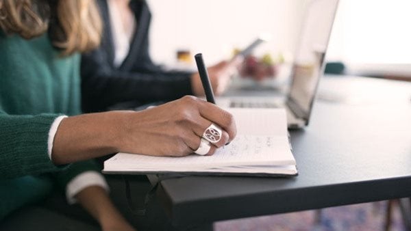 Person skriver för hand i anteckningsbok under en skrivarkurs, med laptop i bakgrunden